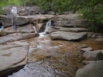 [Lots of horizontal light-colored granite slabs stacked on three different levels. Waterfalls seen in two spots leading to a pool of water in the foreground.]